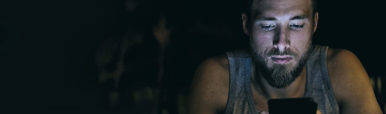 A young man in a tank top sits in his room in the dark reading a text message from his crush, confused about what she's saying.