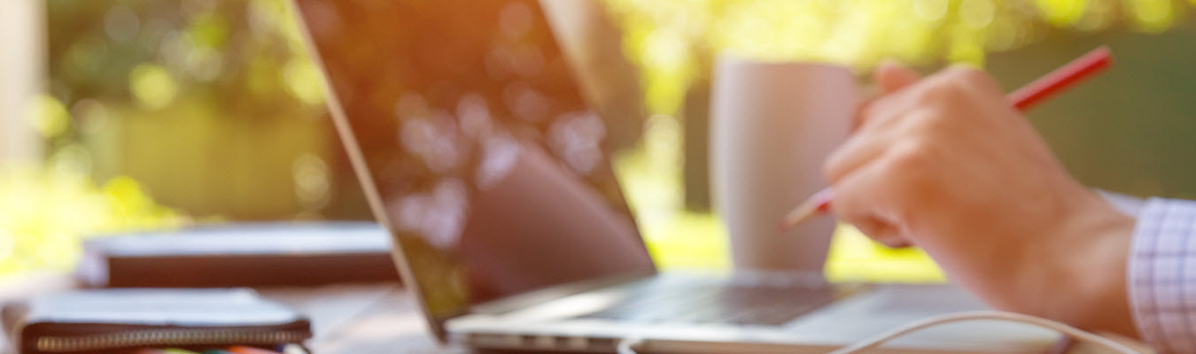 A psychologist sits at his desk outside with his laptop open and pen in hand assembling exciting psychology content to put online. 