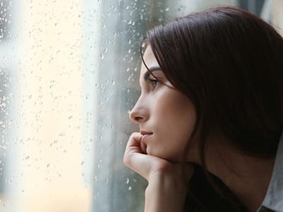 A beautiful young woman anxiously looks out the window while it's raining, constantly thinking something will go wrong. 