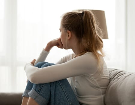 A young depressed woman sits on the couch looking out the window, pensively wondering if things will ever change. 