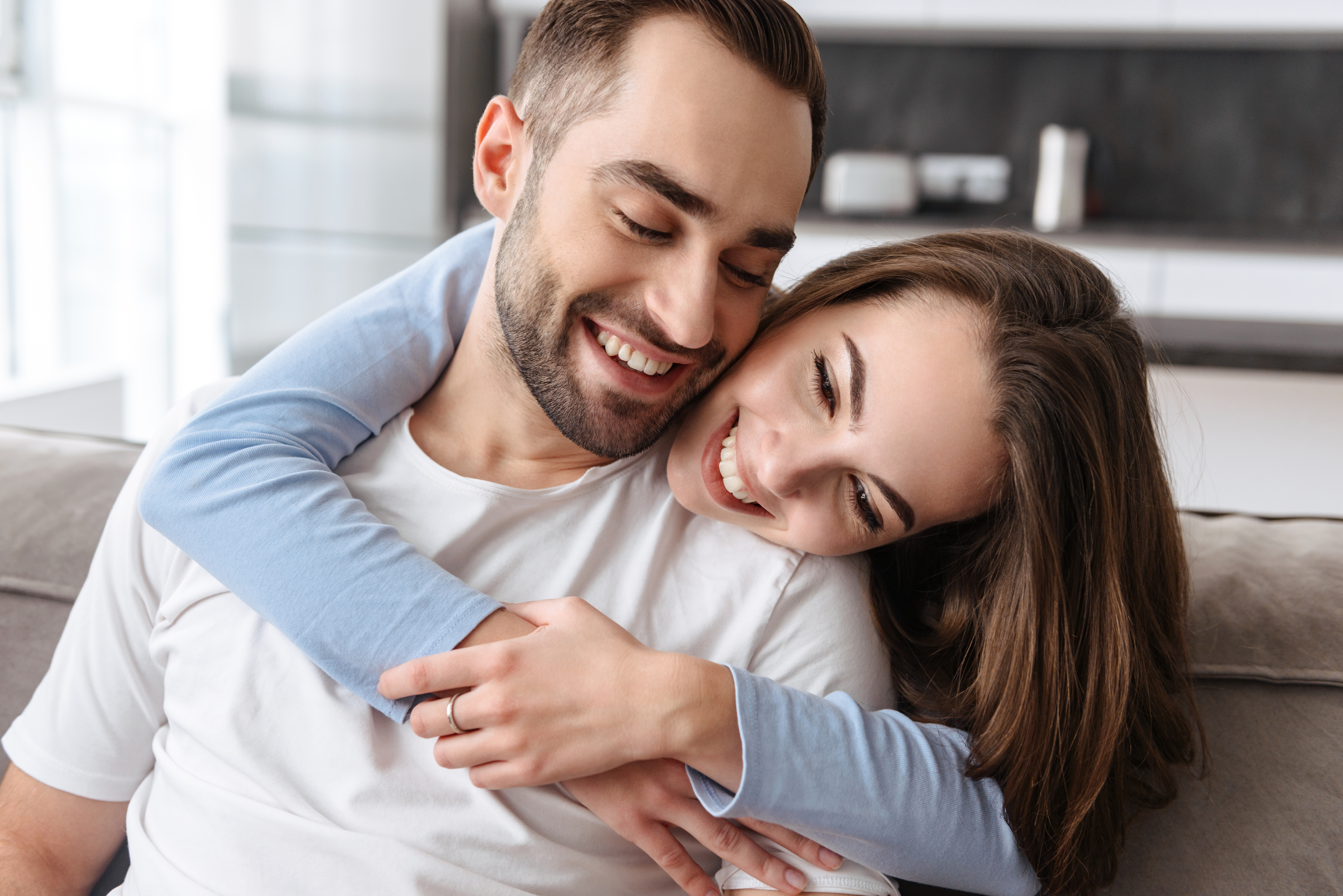 A man sits smiling on the couch as his girlfriend embraces him from behind. They are having fun together.