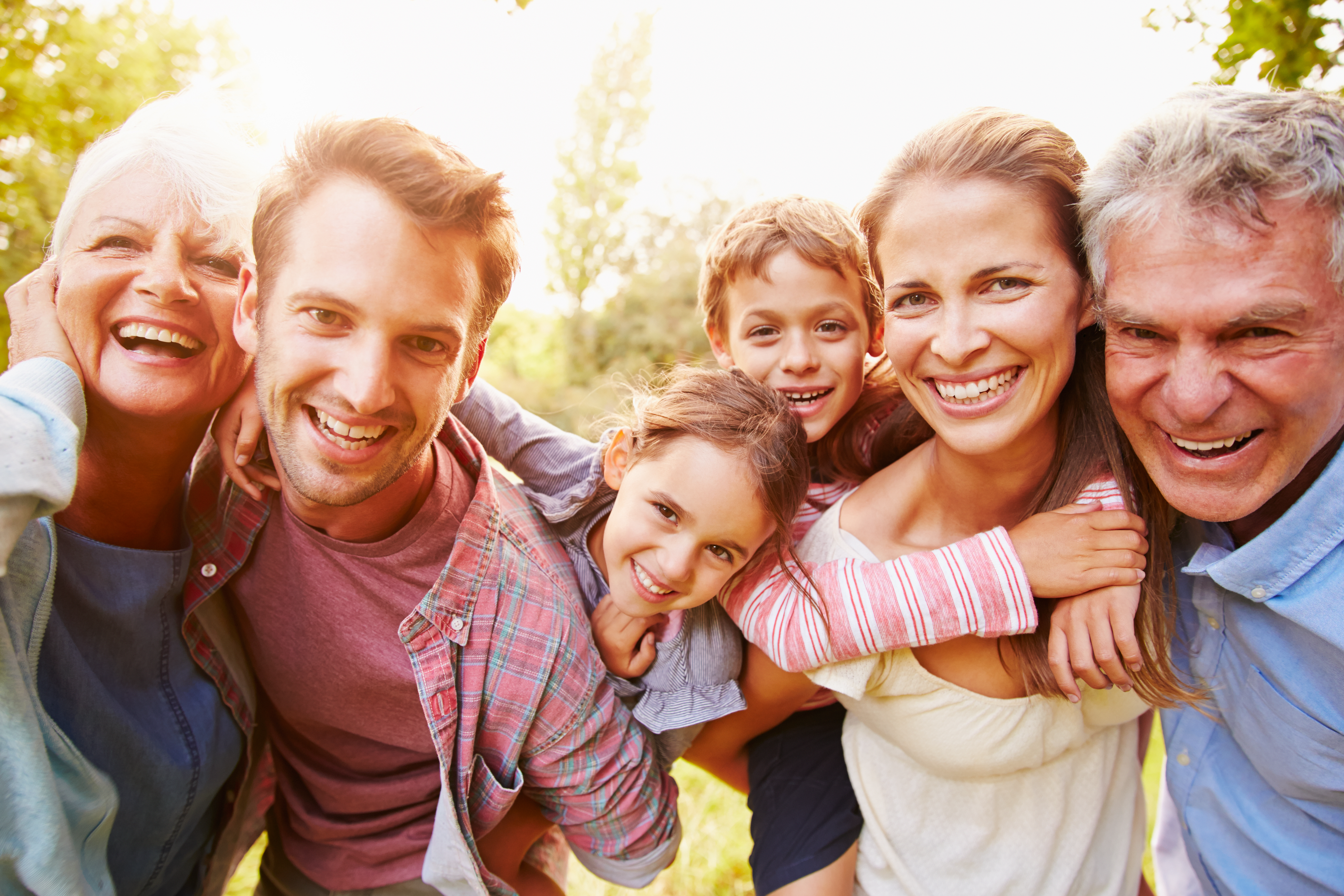 Three generations pose for a fun, loving family picture outdoors with the sun shining behind them.